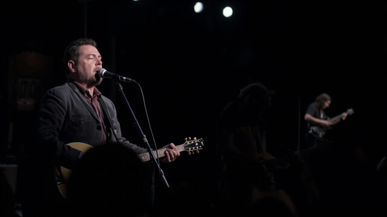 A bluegrass musician performing with a microphone on a dimly lit stage with a guitar player visible in the background