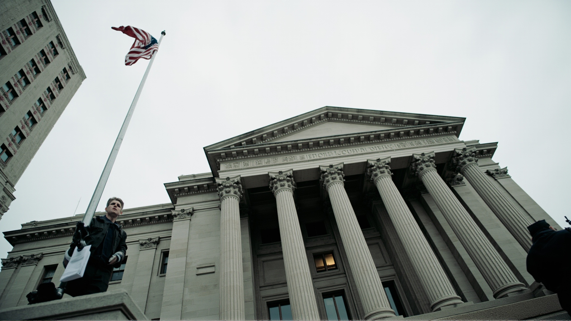 A federal courthouse with classical columns, shot from below, American flag at half-staff, overcast sky, documentary gravitas