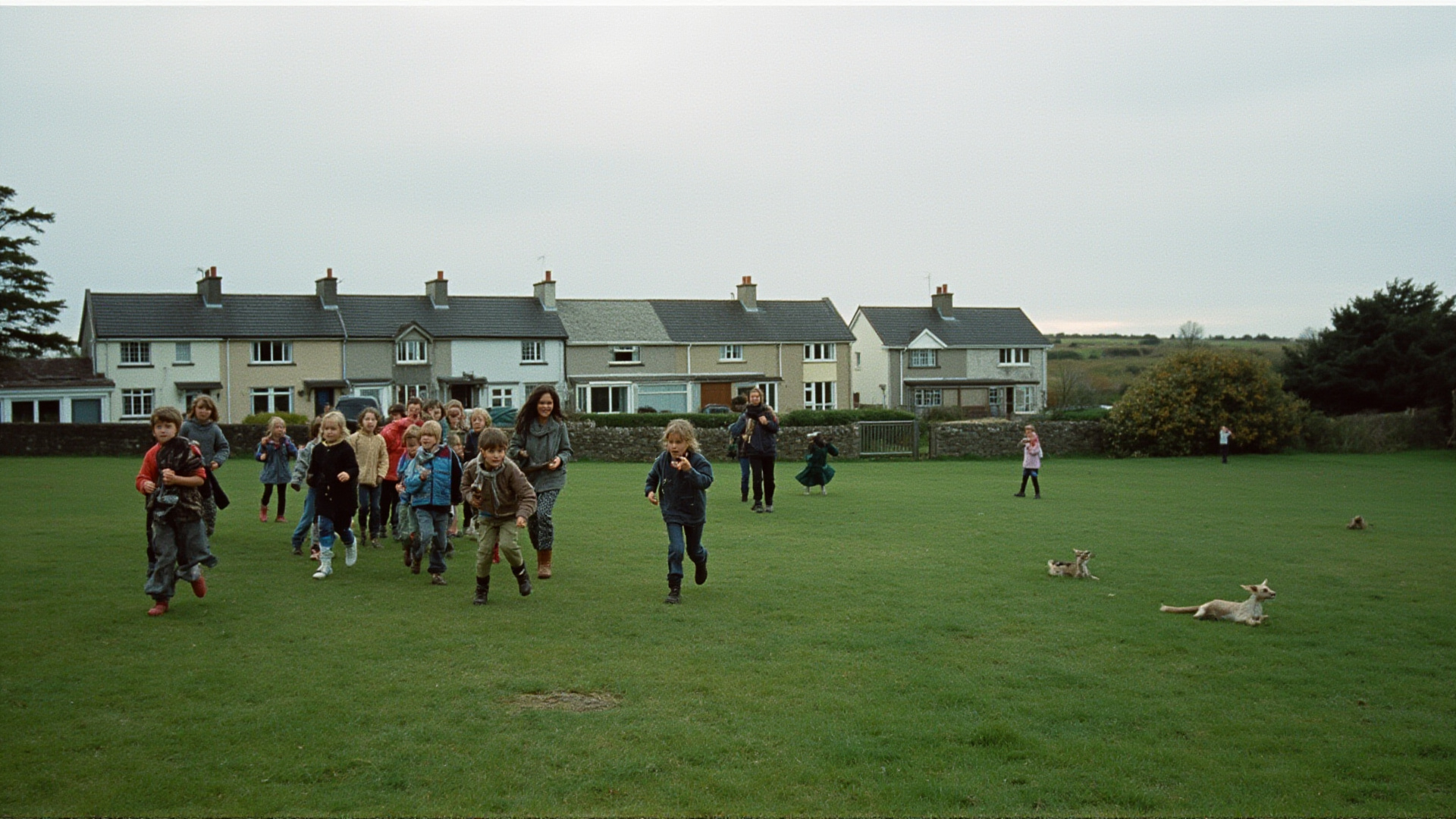 Children playing on a village green in Ireland, overcast sky, no phones visible, a mix of ages running and talking, terraced houses in background