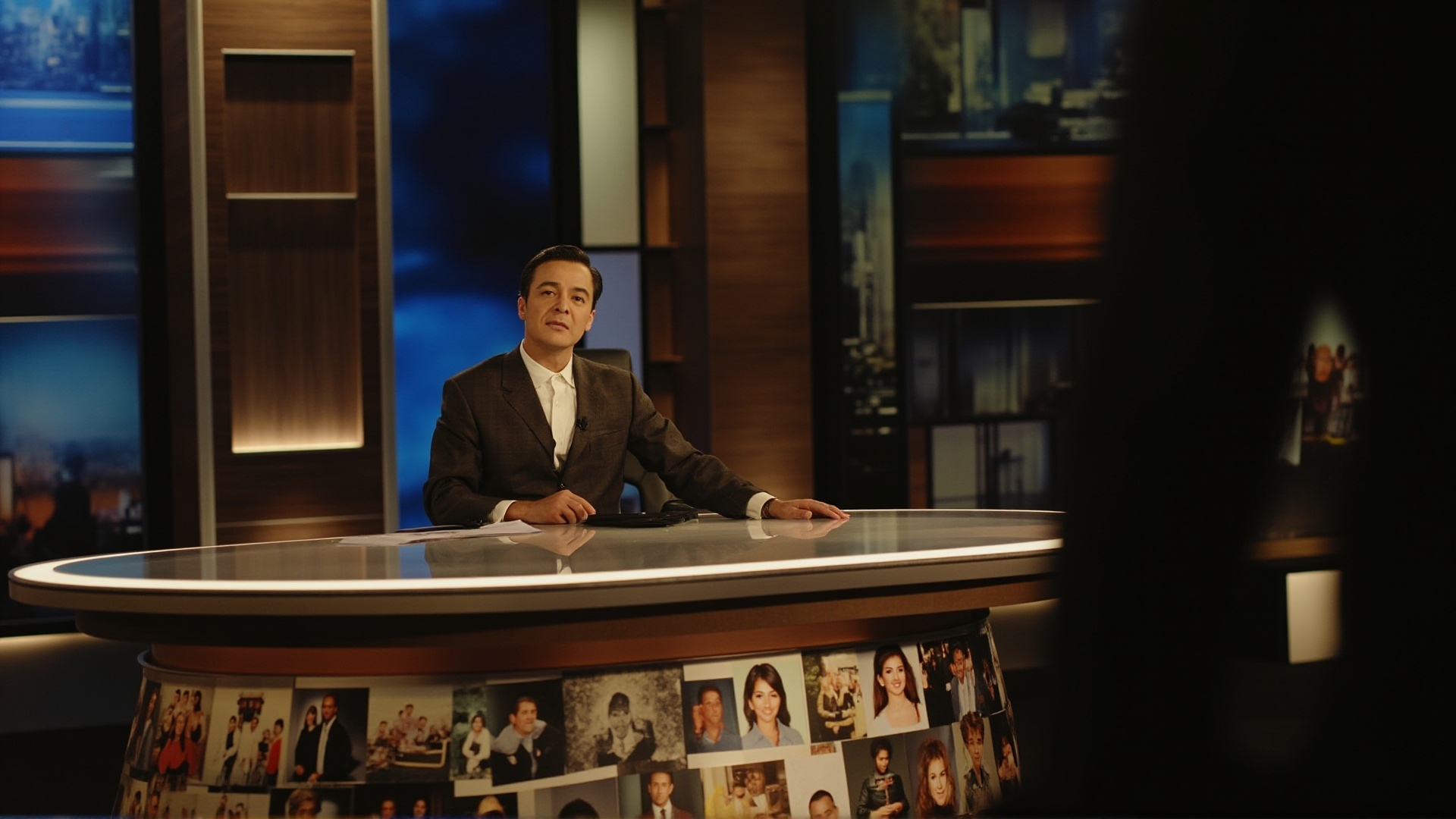 A television studio desk with a single news anchor's chair, warm studio lighting, personal photos visible on the desk edge, intimate broadcast atmosphere