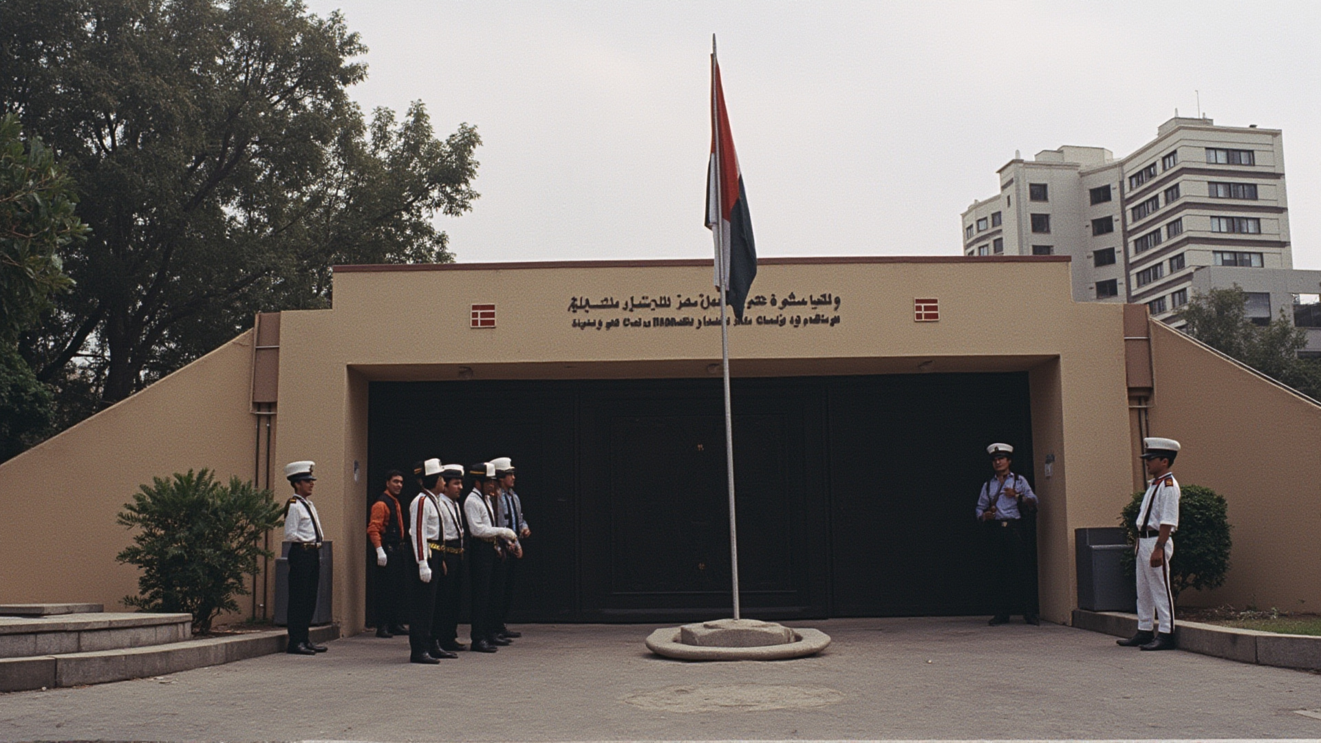 A diplomatic embassy building with a lowered flag, guards at the gate, Beirut urban backdrop, overcast
