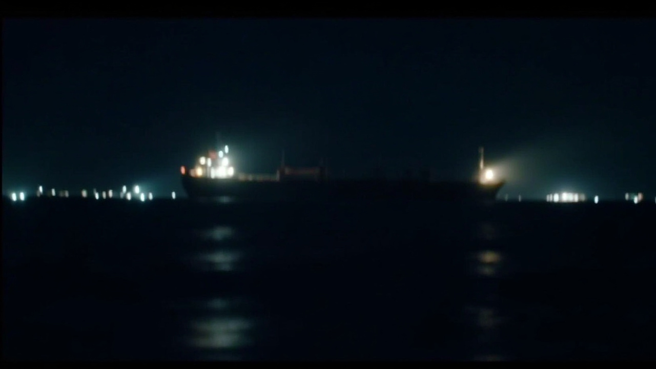A cargo ship anchored in dark waters near the Strait of Hormuz, its lights glowing against a night sky