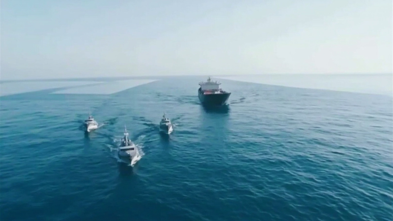 A cargo vessel passing through the Strait of Hormuz flanked by naval patrol boats
