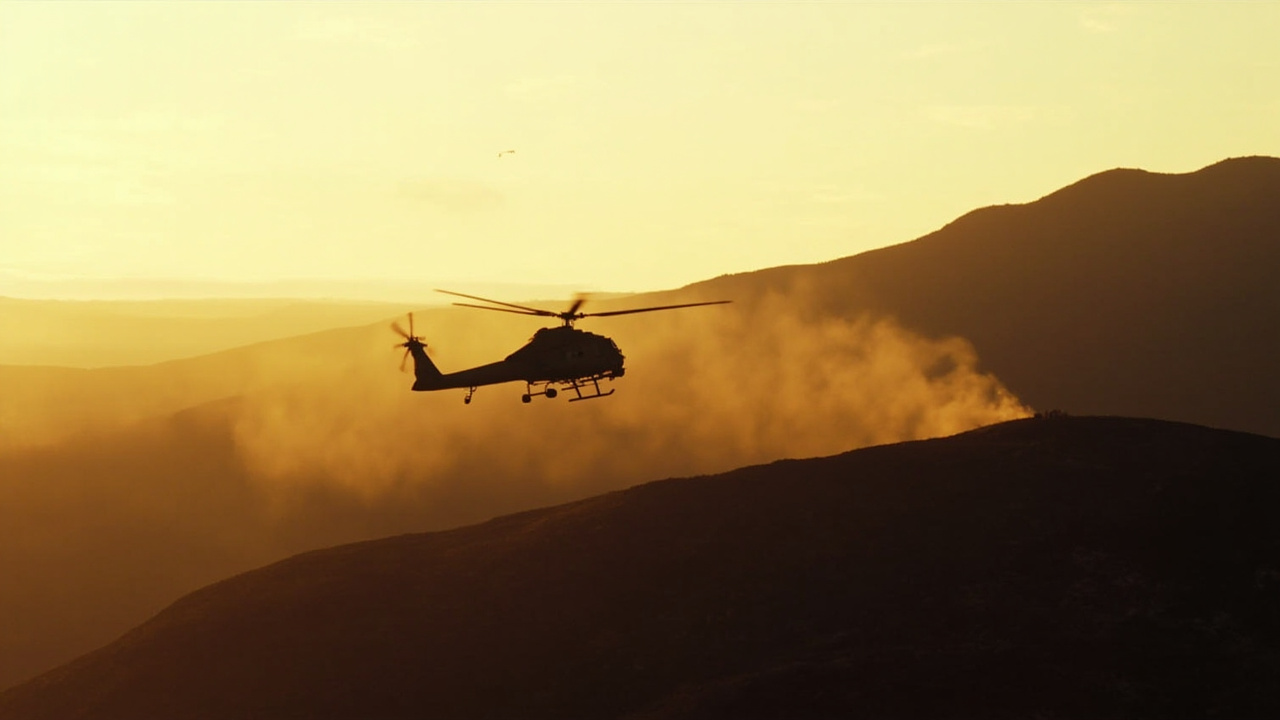 Twilight landscape of rugged Iranian mountain terrain in Kohgiluyeh province with a distant helicopter silhouette against an orange sky, suggesting a search and rescue operation