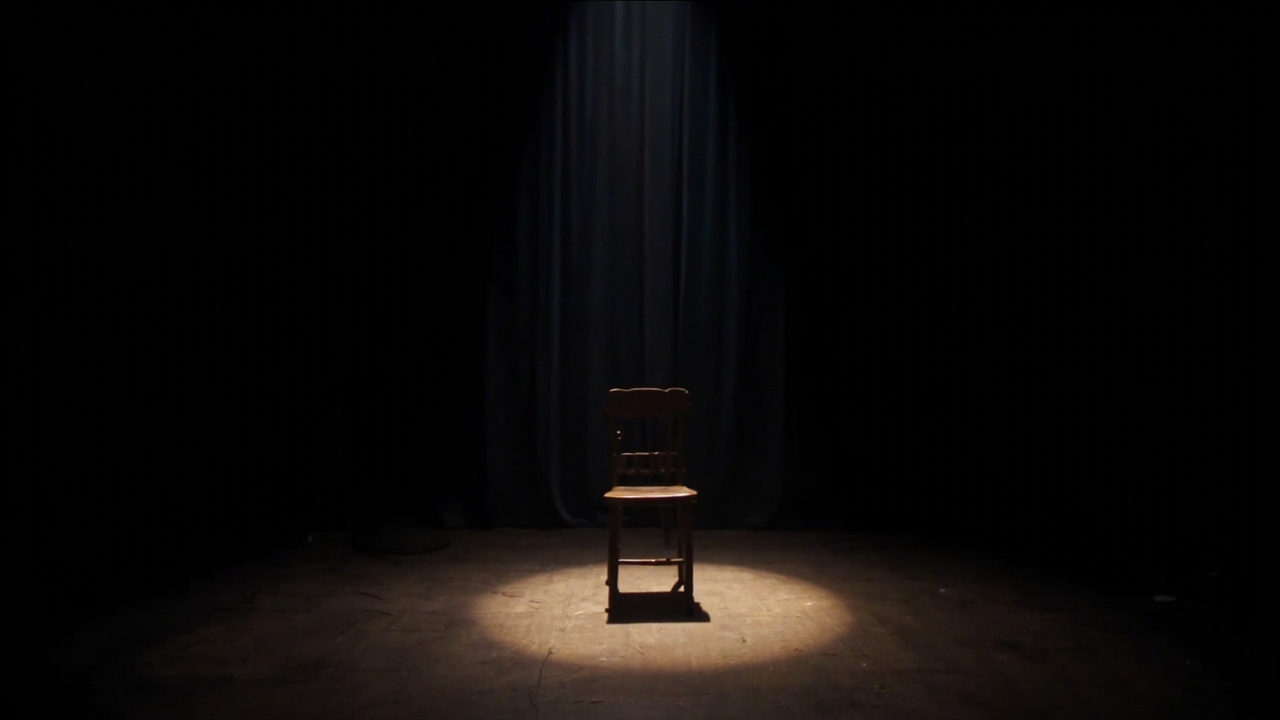 A dimly lit theatre stage with a single spotlight, an empty chair center stage, evoking the absence of a performer
