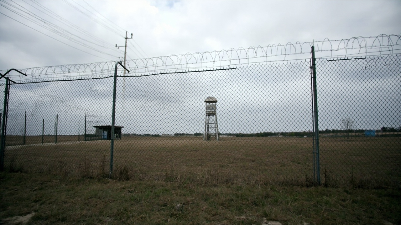 The exterior of a rural correctional facility with a chainlink fence and guard tower visible under an overcast sky