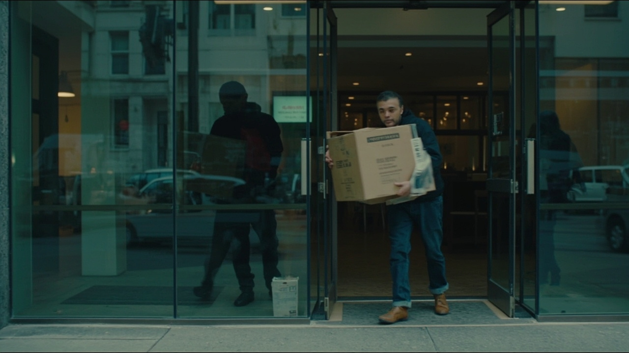 Former tech worker carrying a box of belongings from an office building, other employees visible in background