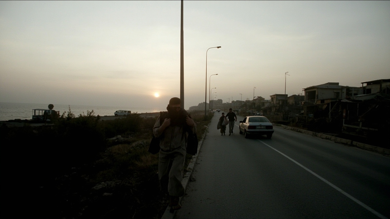 A coastal road in southern Lebanon at dusk, a few cars stopped, families carrying bags on foot toward villages with damaged rooftops