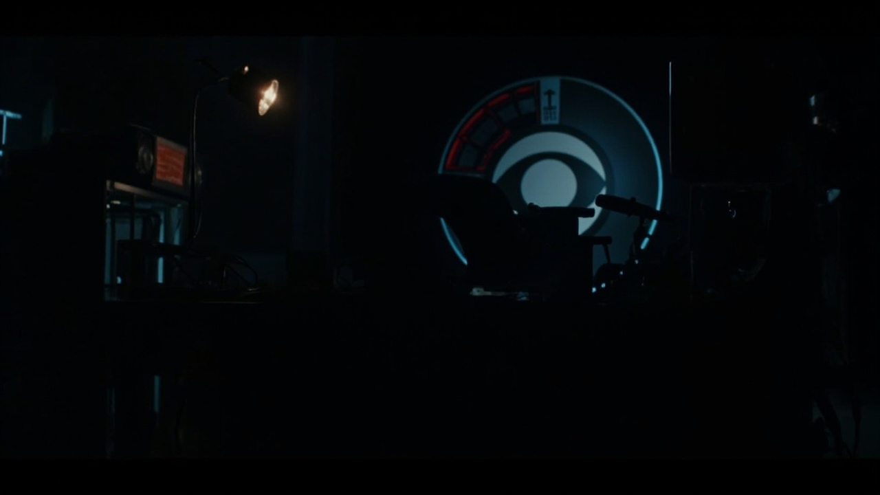 A dim CBS News Radio studio at night, empty chair behind the microphone, broadcast clock at the top of the hour