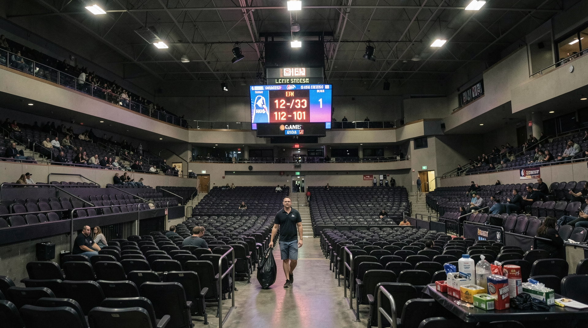 Little Caesars Arena after Game 1 Sunday night, wide shot from the upper bowl, the lights up, confetti unused on the mezzanine table, a single custodial worker walking the aisle, the scoreboard still showing the final.