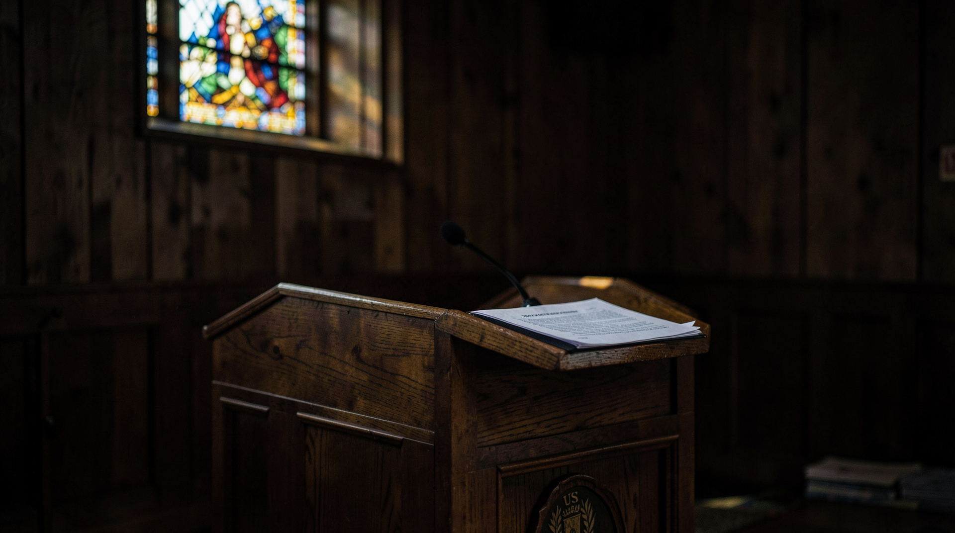 A USCCB podium bearing the Conference seal, empty at the moment, a folder of printed doctrinal statements on the lectern, stained-glass light falling across the far wall.