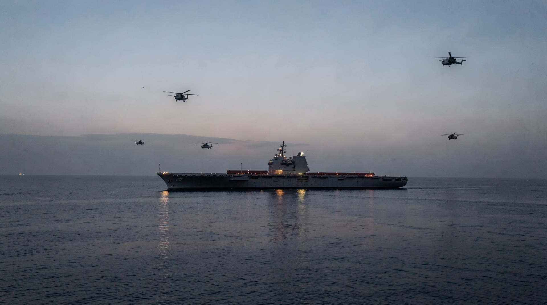 A U.S. carrier strike group at dusk in the Gulf of Oman with a stationary tanker silhouetted in the middle distance.