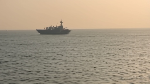 An oil tanker silhouetted against a hazy dawn near the Strait of Hormuz, with a small naval vessel visible in the distance.