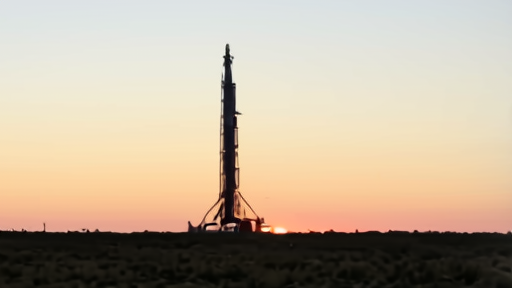 A Falcon 9 standing on a launch pad at twilight, ground-lighting on the strongback.