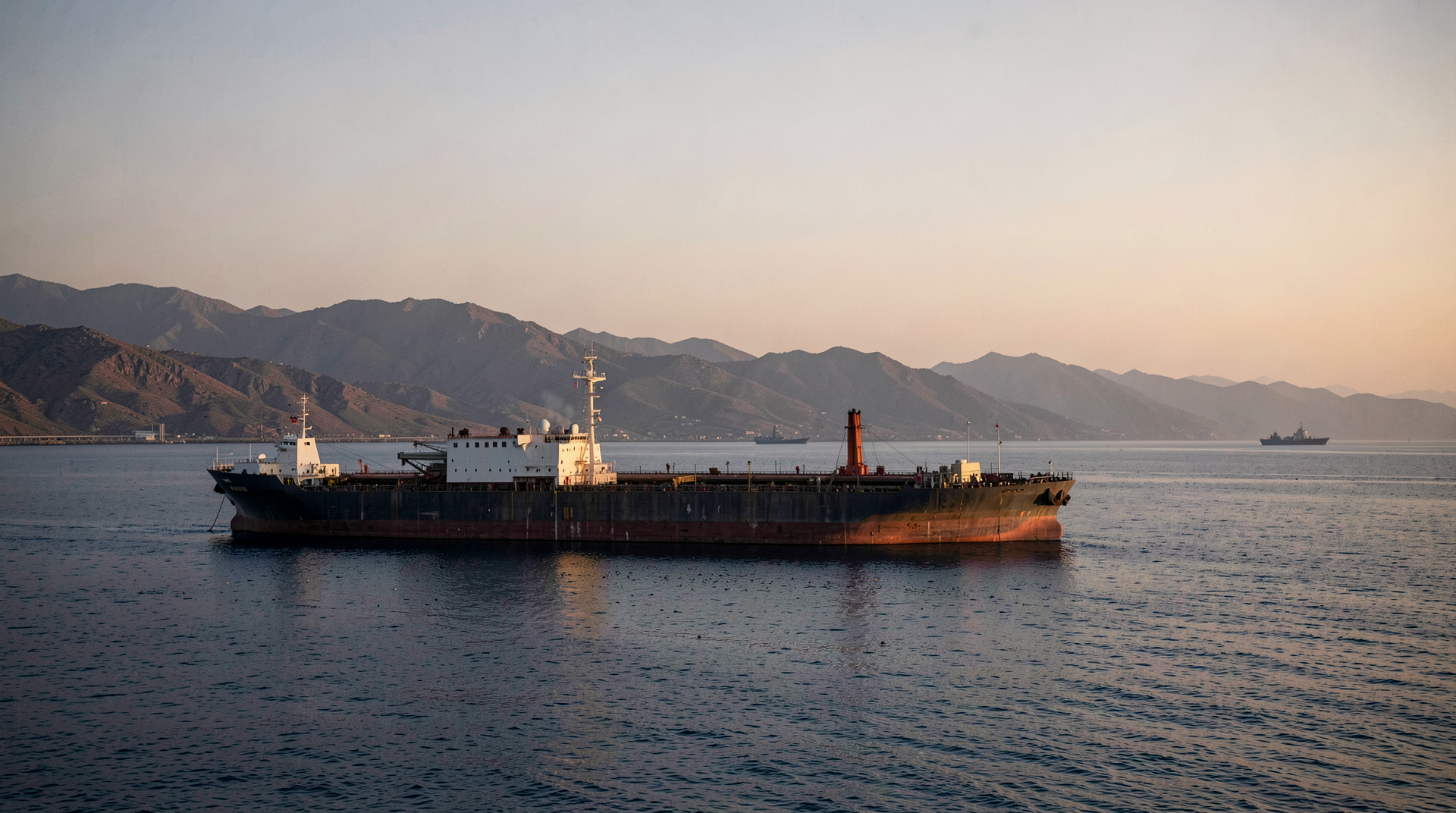An oil tanker silhouetted against a dawn sky in the Strait of Hormuz with Omani mountains visible behind.