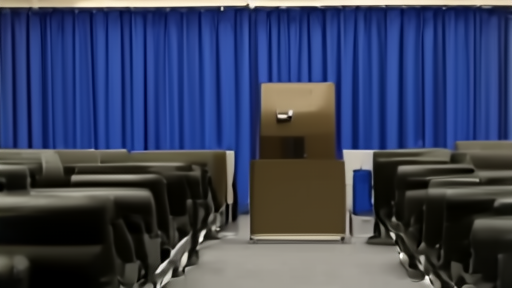 The Pentagon press briefing room at the end of a workday, the lectern empty, blue curtain hanging behind it, rows of empty press seats facing forward.