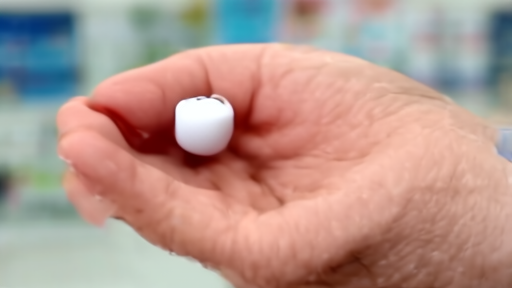 A hand holding a single round Foundayo tablet against the blurred backdrop of a pharmacy counter.