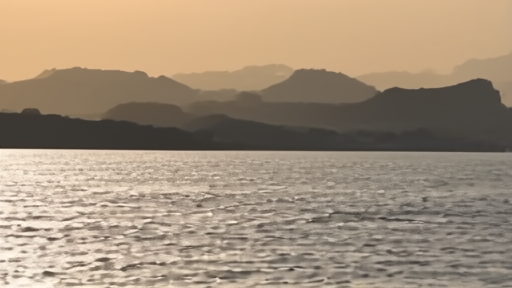 Large red-and-grey LNG tanker silhouetted at sunset against the low Iranian Hormuz coastline, calm water, escort vessel in the wake
