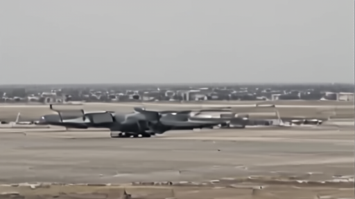 Overhead satellite-style frame of a tarmac near a hangar at a military airbase, the silhouette of a four-engine C-130-type transport plane visible at the edge of a parking apron.