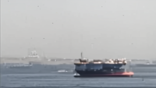 A tanker moves through a narrow strait while a tracking screen glows nearby