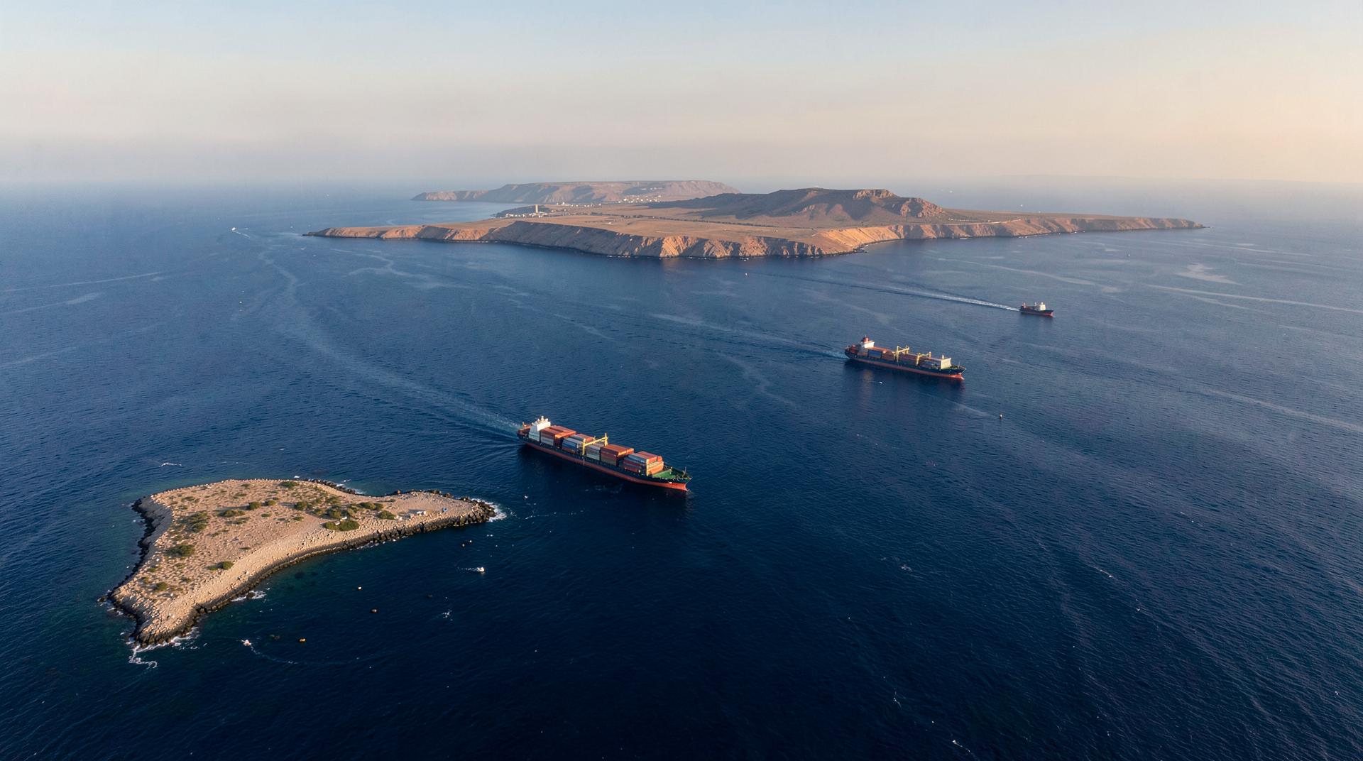 Aerial wide-angle of the Strait of Hormuz, Hormuz Island in foreground with Qeshm Island in middle distance, two container ships transiting the channel under late-afternoon light.