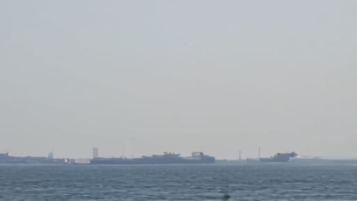 Tankers waiting offshore near a hazy shipping lane with naval vessels in the distance