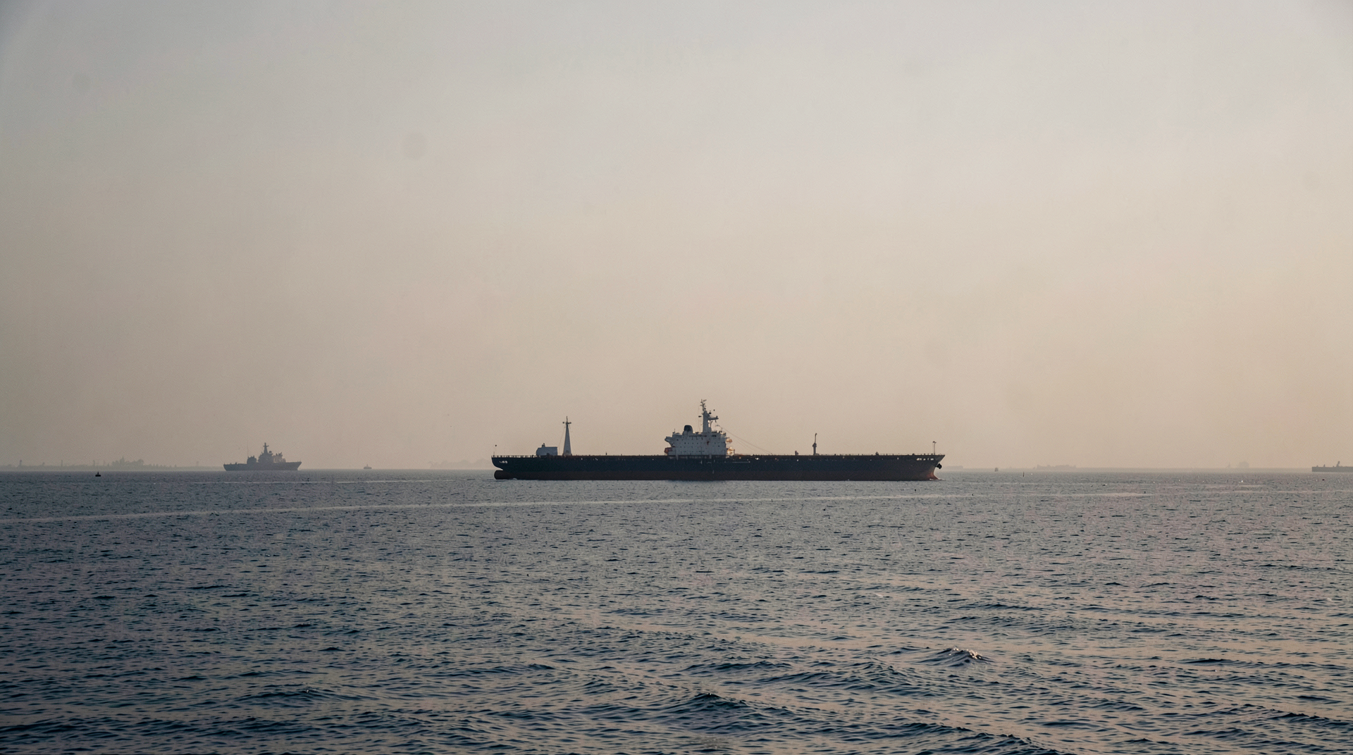 Strait of Hormuz at dawn with a single tanker silhouetted against the Iranian coast and a naval vessel on the horizon