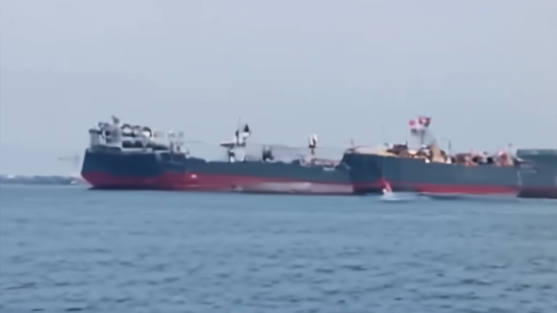 Line of commercial vessels anchored in open water with military escort visible in distance