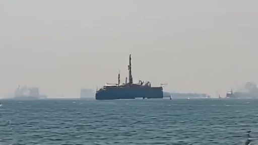 An oil tanker seen from a distance in the Strait of Hormuz with gray naval vessels flanking both sides