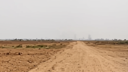 A dusty road in a rural African landscape with smoke rising from a distant settlement against a hazy sky